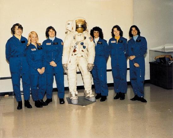 Six women in astronaut flight suits pose with a spacesuit